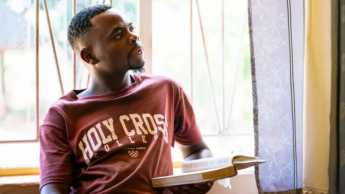 Ein Mann in einem Hemd des Holy Cross College sitzt an einem Fenster, hält ein aufgeschlagenes Buch in der Hand und schaut zur Seite.
