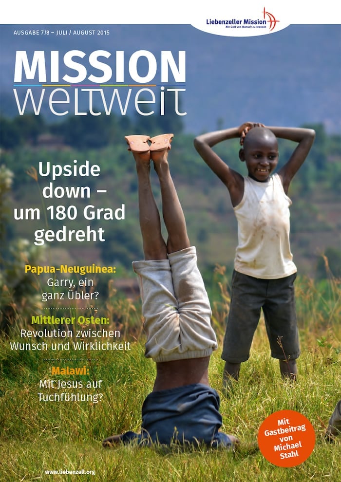 Zwei lächelnde Kinder spielen draußen; eines steht und das andere macht einen Handstand auf einer Wiese mit Bergen im Hintergrund.