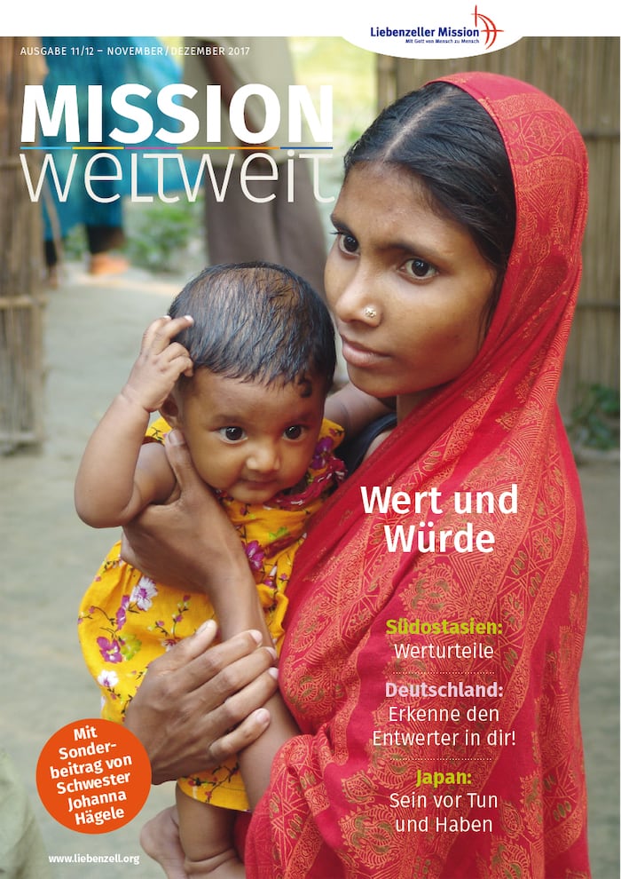 Eine Frau mit rotem Schal hält ein Baby in einem gelben Outfit, beide schauen auf einem Dorfweg in die Kamera.