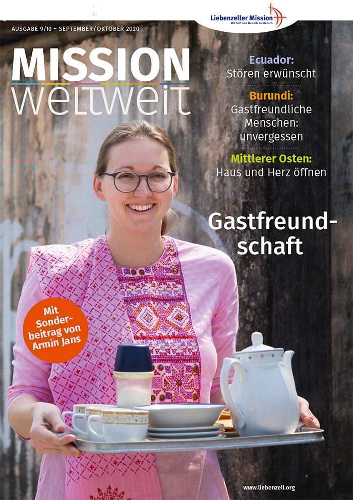 Eine lächelnde Frau hält ein Tablett mit Tee im Freien, sie trägt eine Brille und ein rosa besticktes Kleid.