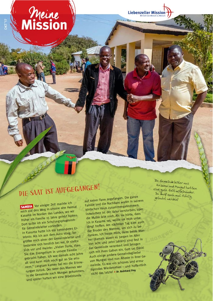 Vier lächelnde Männer stehen im Freien im Sonnenlicht zusammen und halten sich an den Händen, mit einem Gebäude und anderen Menschen im Hintergrund.