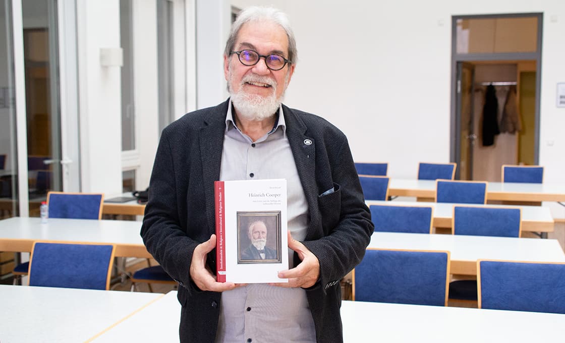 Ein lächelnder Mann mit Brille hält ein Buch mit einem Porträt auf dem Einband in einem Klassenzimmer mit blauen Stühlen und weißen Wänden.