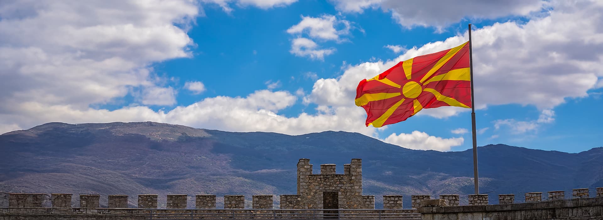Die mazedonische Flagge weht über einer steinernen Festung mit Bergen und einem teilweise bewölkten Himmel im Hintergrund.