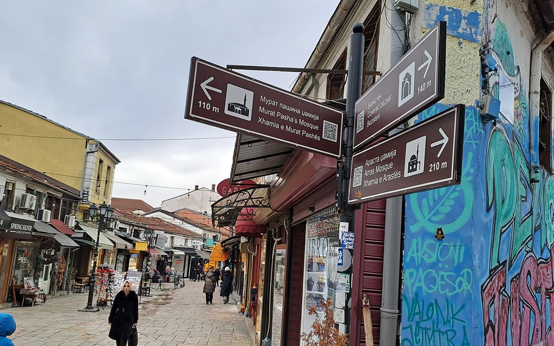 Straßenszene mit Spaziergängern, Geschäften, Graffiti und braunen Touristenschildern, die den Weg zu örtlichen Sehenswürdigkeiten weisen.