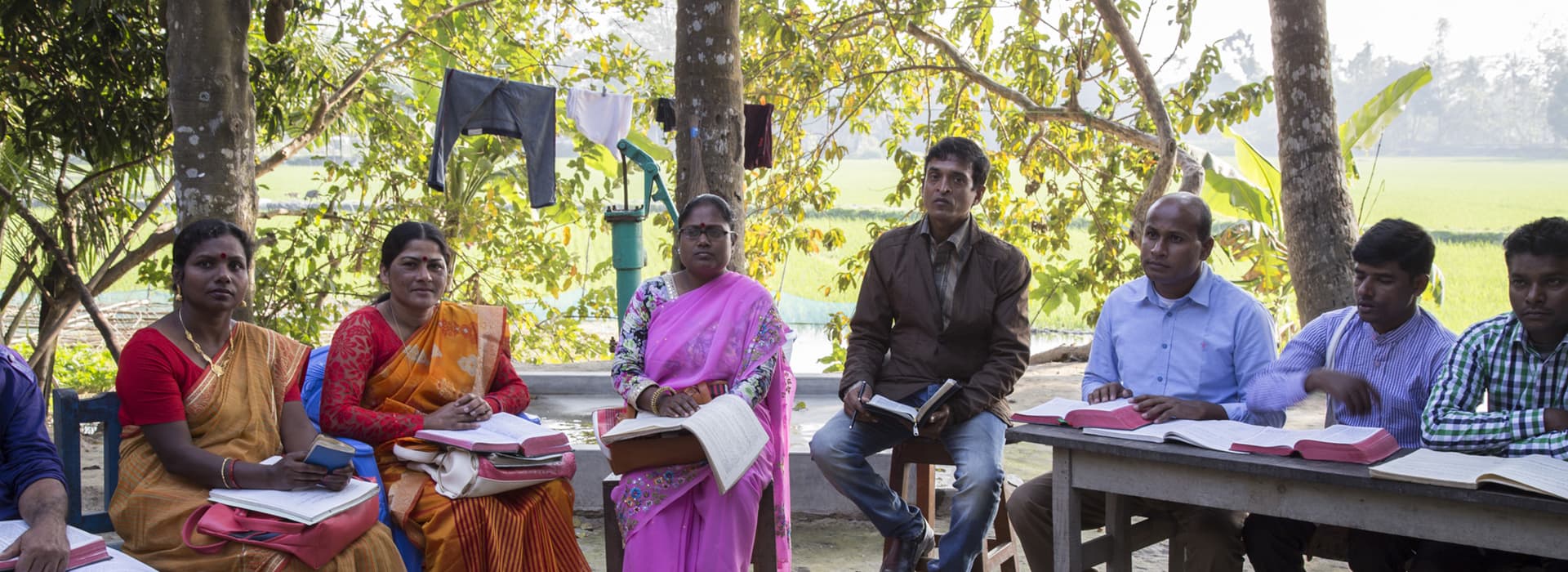 Eine Gruppe von Menschen sitzt im Freien an Tischen unter Bäumen, hält Bücher in der Hand und hält eine Besprechung oder eine Studiensitzung ab.