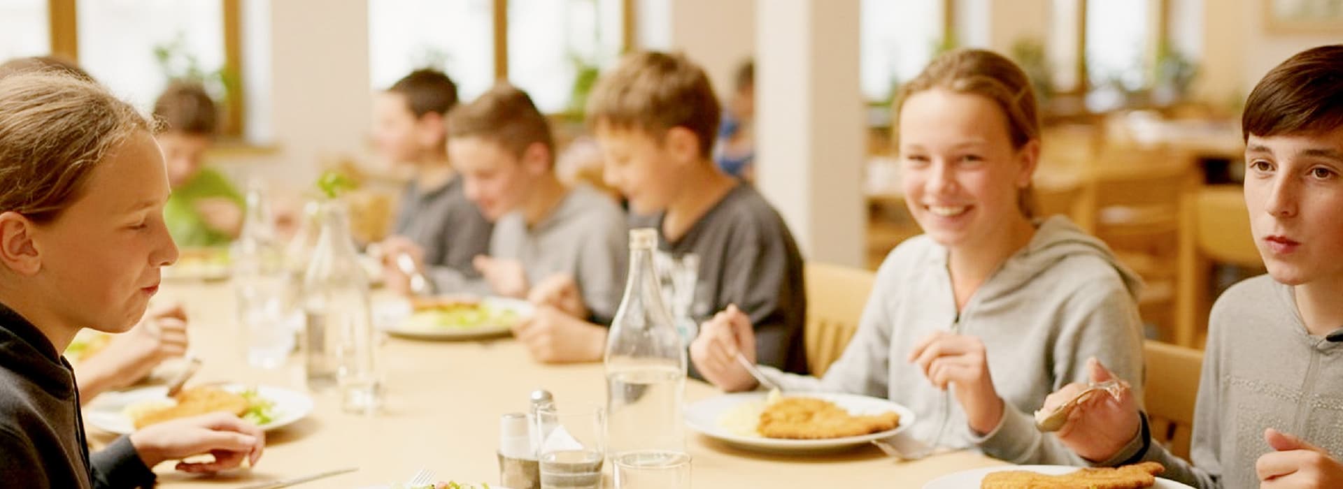 Eine Gruppe lächelnder Schüler sitzt an einem Tisch in der Cafeteria und isst gemeinsam in einem hellen, luftigen Raum zu Mittag.