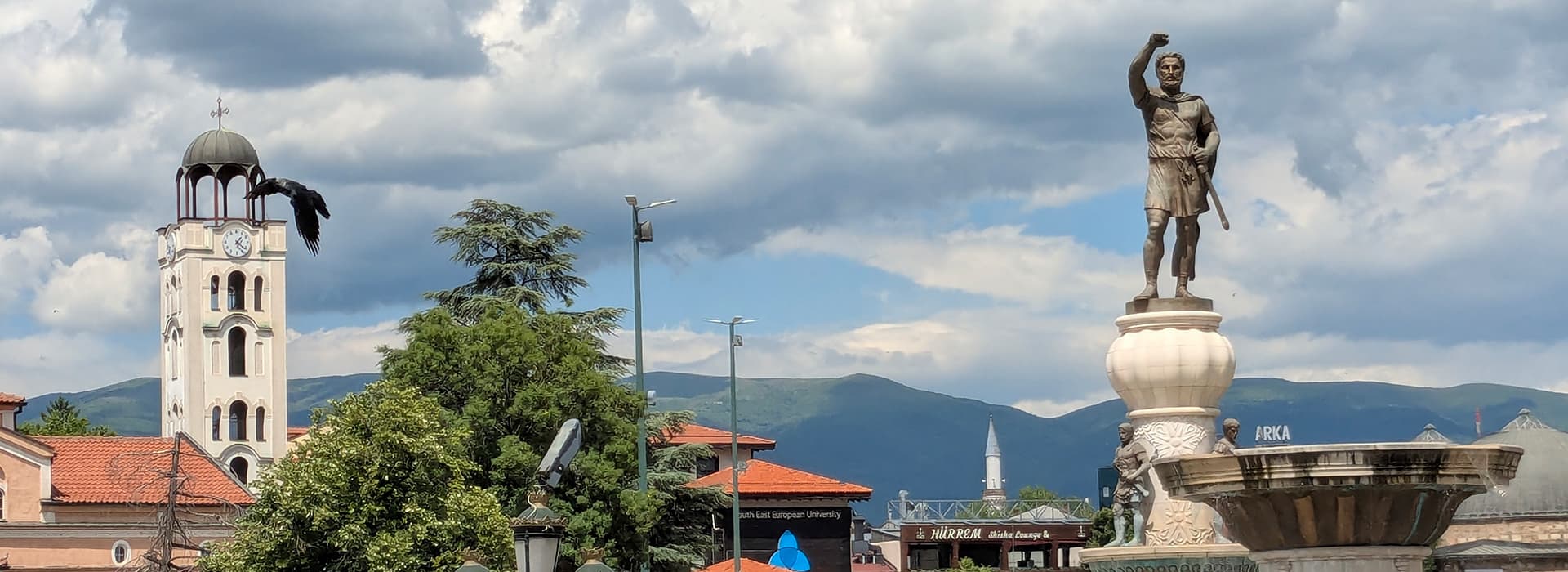Eine Statue mit erhobenem Arm steht an einem Brunnen; im Hintergrund ist ein Kirchturm unter einem bewölkten Himmel zu sehen.