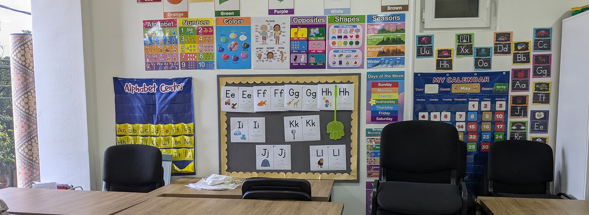 Eine farbenfrohe Klassenzimmerwand mit Lernplakaten, eine Alphabettafel und mehrere leere schwarze Stühle an Holzpulten.
