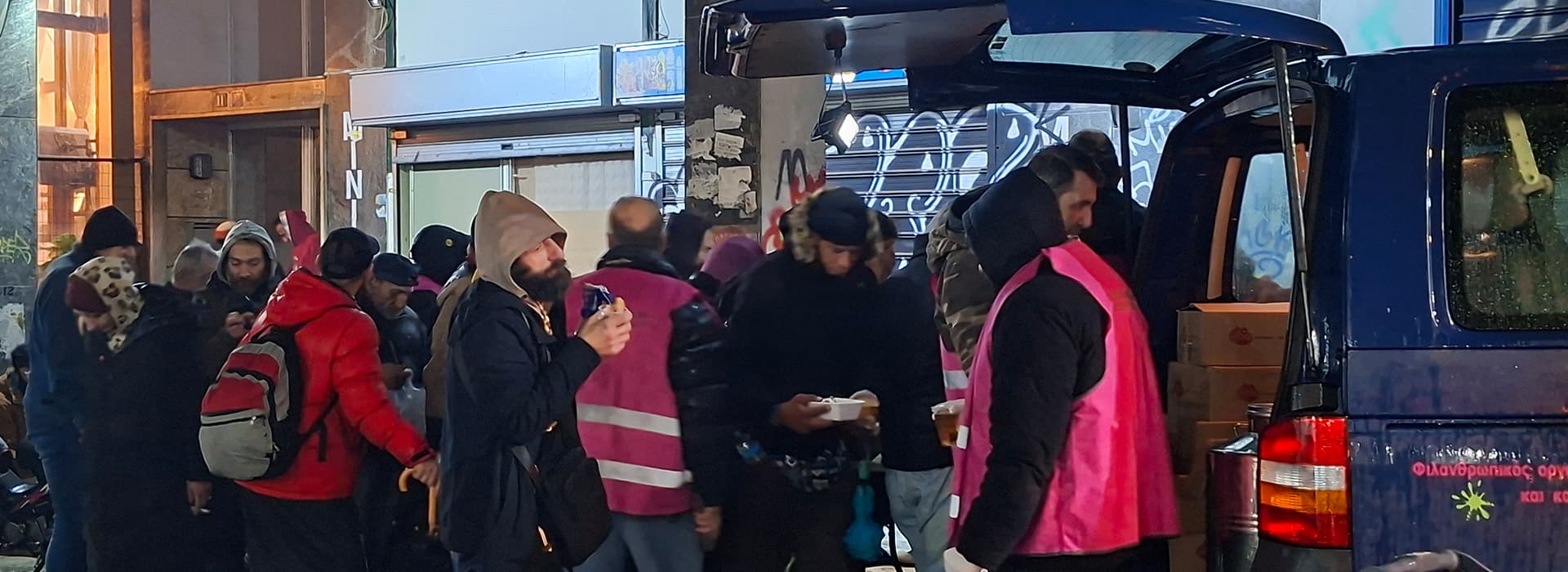 Freiwillige Helfer in rosafarbenen Westen verteilen Lebensmittel und Getränke an Menschen, die nachts in einer Schlange vor einem blauen Lieferwagen stehen.