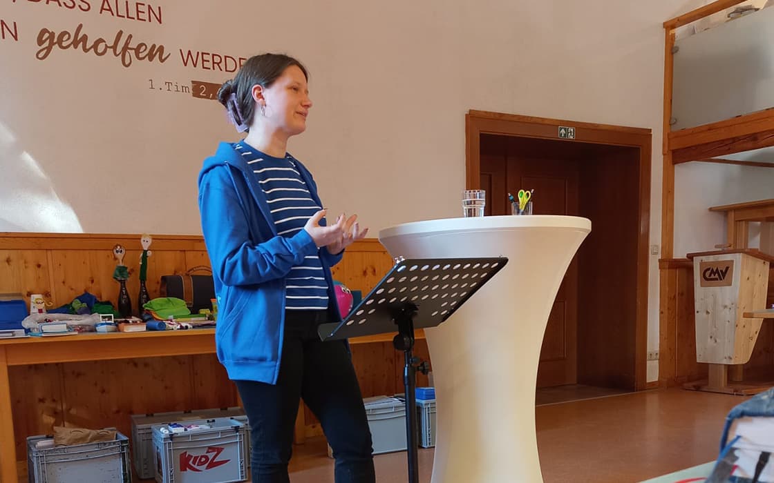 Eine Frau in einem blauen Kapuzenpulli spricht an einem Podium in einem Raum mit Holzwänden und verschiedenen Materialien auf einem Tisch hinter ihr.