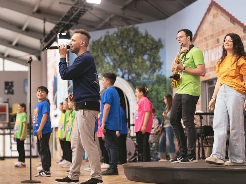 Ein Mann singt in ein Mikrofon, während Kinder in bunten Hemden und ein Musiker auf der Bühne in einem Innenraum stehen.