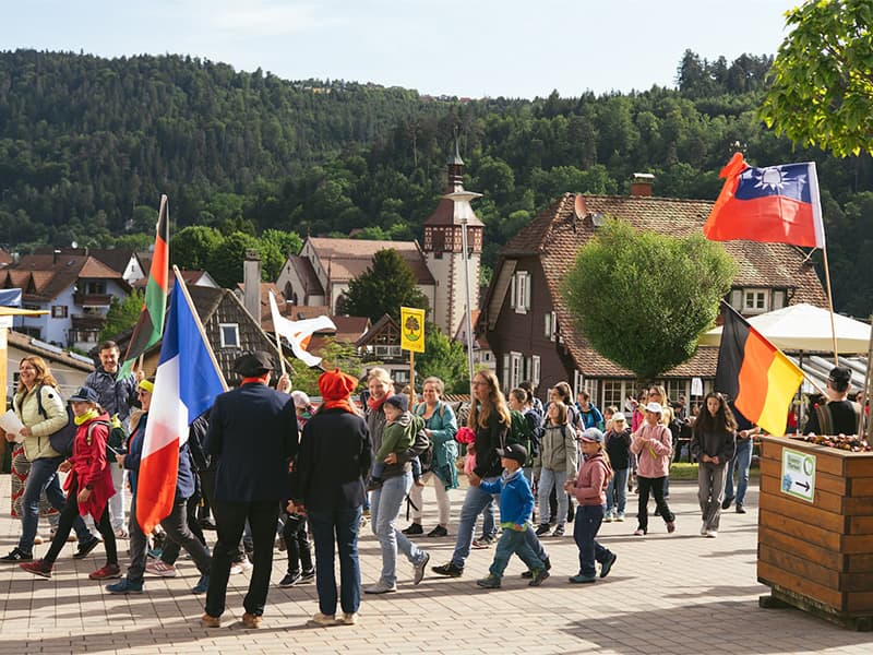 Eine Gruppe von Menschen mit Fahnen geht im Freien in einem Dorf mit grünen Hügeln und Häusern im Hintergrund spazieren.