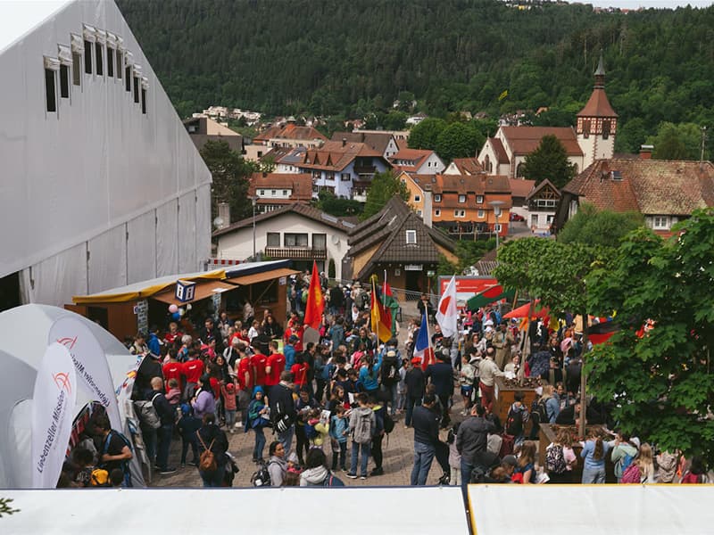 Eine große Menschenmenge versammelt sich bei einer Veranstaltung im Freien in einer Stadt mit Zelten, Fahnen und bewaldeten Hügeln im Hintergrund.