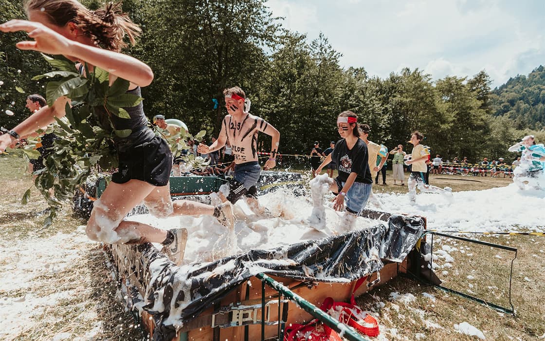 An einem sonnigen Tag rennen Menschen in Kostümen und mit Gesichtsbemalung durch einen mit Schaumstoff gefüllten Hindernisparcours im Freien.