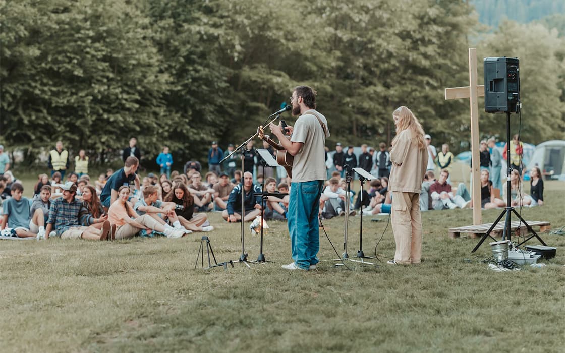 Ein Mann und eine Frau spielen Musik auf einer grasbewachsenen Bühne im Freien vor einem sitzenden Publikum in der Nähe von Bäumen und einem Holzkreuz.