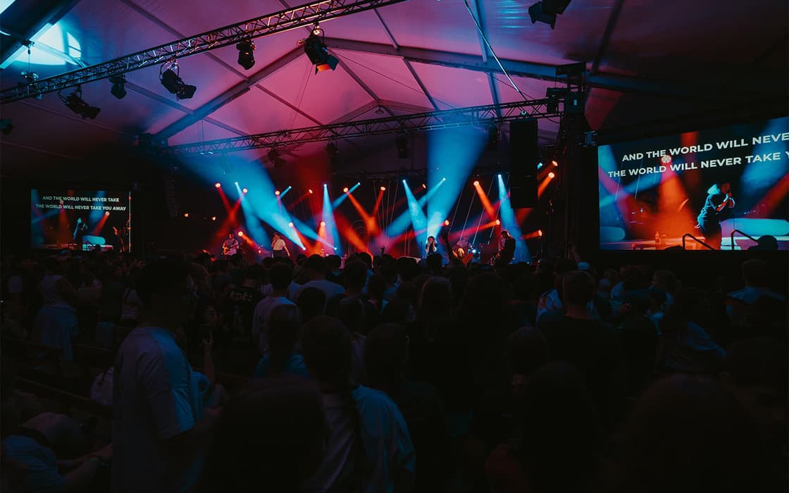 Menschenmenge bei einem Live-Konzert in einer Halle mit bunten Bühnenlichtern und auf eine Leinwand projizierten Liedtexten.