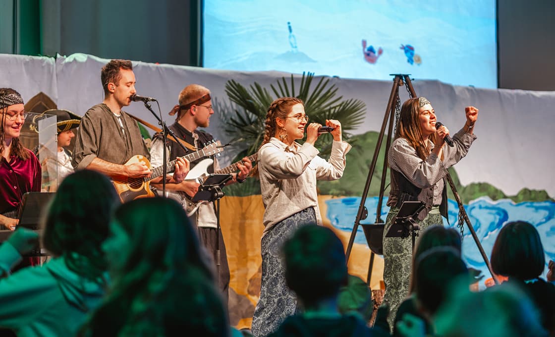 Eine Gruppe von Menschen spielt auf der Bühne vor einem Publikum vor einer farbenfrohen Strandkulisse Musik.