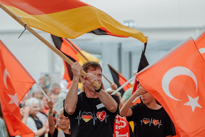 Menschen schwenken deutsche und türkische Fahnen bei einer Indoor-Veranstaltung und tragen Hemden mit deutschen und türkischen Herzsymbolen.