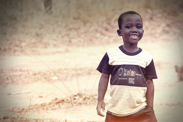 Ein lächelnder Junge in einem gestreiften T-Shirt steht im Freien auf einem unbefestigten Boden mit trockenem Gras im Hintergrund.