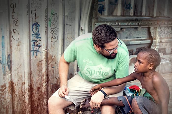 Ein Mann in einem grünen Hemd tröstet einen jungen, hemdlosen Jungen, der draußen vor einer mit Graffiti beschmierten Metallwand sitzt.