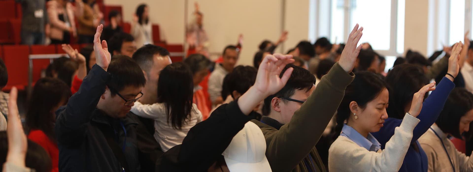 Eine Gruppe von Menschen mit erhobenen Händen nimmt an einer Versammlung in einem hellen Raum mit roten Sitzen teil.