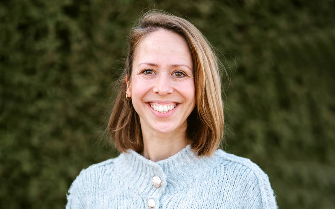 Eine lächelnde Frau mit schulterlangem Haar in einem leichten Strickpullover steht draußen vor einer Grünanlage.