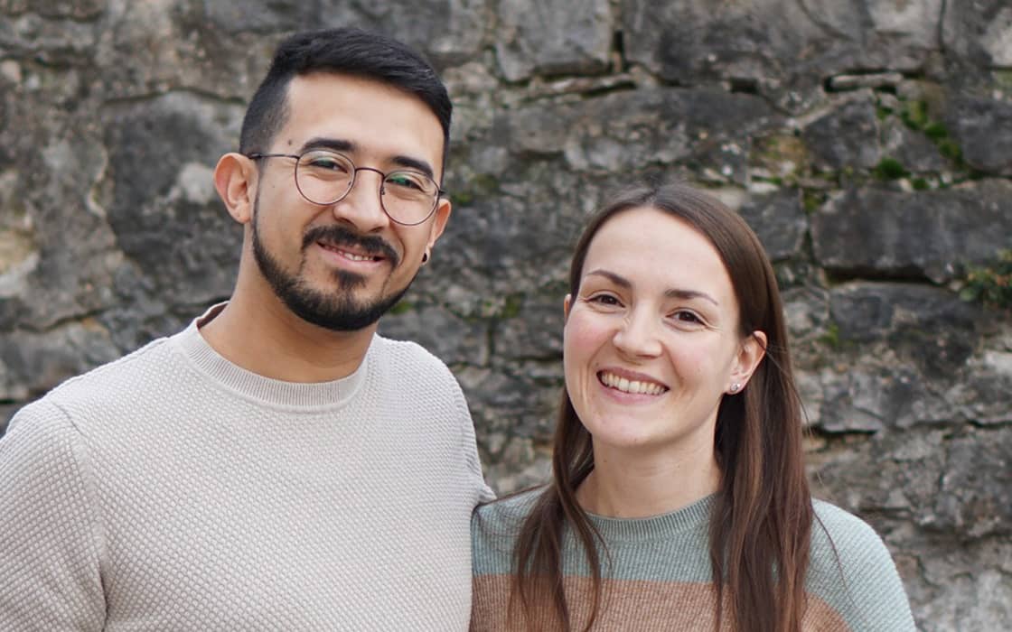 Ein lächelnder Mann und eine lächelnde Frau stehen dicht nebeneinander vor einer Steinmauer.