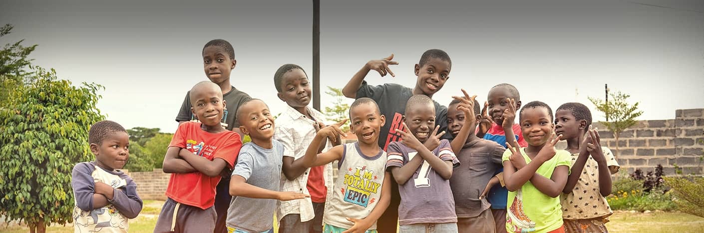 Eine Gruppe lächelnder Kinder posiert spielerisch im Freien vor Bäumen und einer Backsteinmauer.