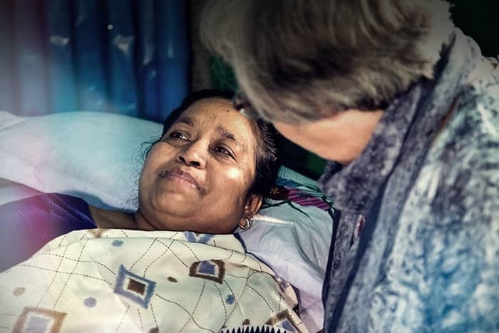 Eine Frau liegt im Bett unter einer Decke, während eine andere Person neben ihr sitzt und sie tröstet und unterstützt.