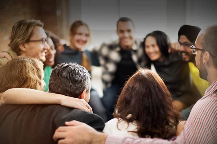 Eine Gruppe von Menschen sitzt eng im Kreis, lächelnd und mit den Armen um die Schultern der anderen.