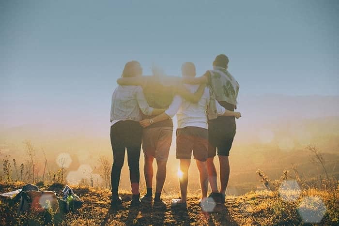 Vier Menschen stehen bei Sonnenuntergang im Freien dicht beieinander, die Arme umeinander gelegt, mit Blick auf die Sonne und die Landschaft.