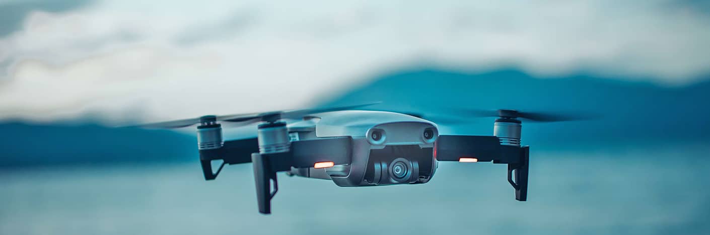 Eine kleine Drohne mit einer Kamera schwebt in der Luft, mit verschwommenen Bergen und Wasser im Hintergrund.