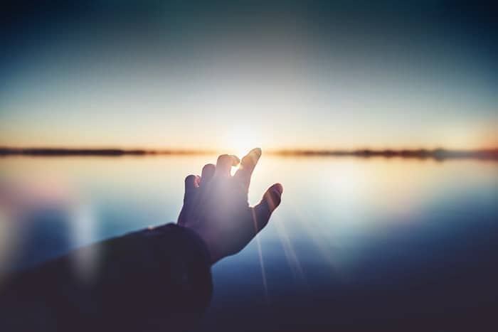 Eine Hand, die sich über einem ruhigen, spiegelnden Gewässer der untergehenden Sonne entgegenstreckt.