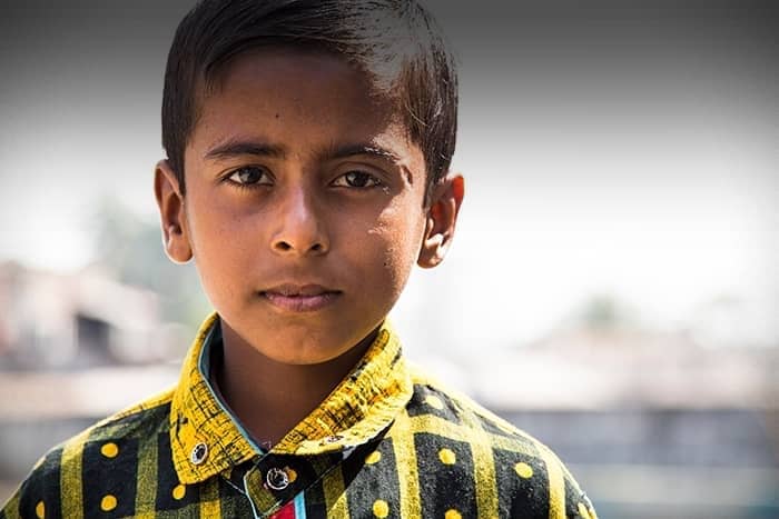 Ein Junge mit kurzen dunklen Haaren und einem gelbkarierten Hemd blickt mit neutraler Miene in die Kamera.