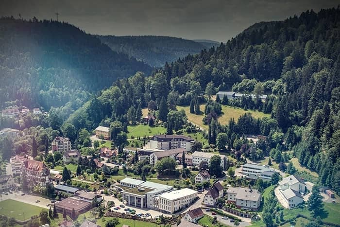 Eine kleine Stadt, eingebettet in ein grünes, bewaldetes Tal, umgeben von Hügeln unter einem wolkenverhangenen Himmel.