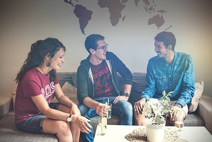 Drei Personen sitzen auf einer Couch, lächeln und unterhalten sich, während sie Getränke in der Hand halten, mit einer Weltkarte an der Wand hinter ihnen.