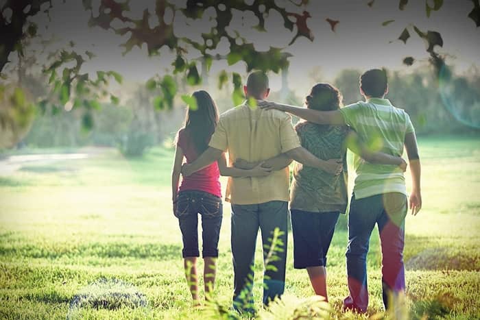 Vier Personen stehen im Freien, die Arme umeinander gelegt, mit dem Gesicht zur Seite, und blicken auf eine sonnenbeschienene Wiese.