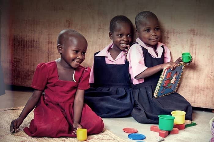 Drei lächelnde junge Mädchen sitzen in Uniformen auf dem Boden und spielen mit buntem Spielzeug und einem Buch.