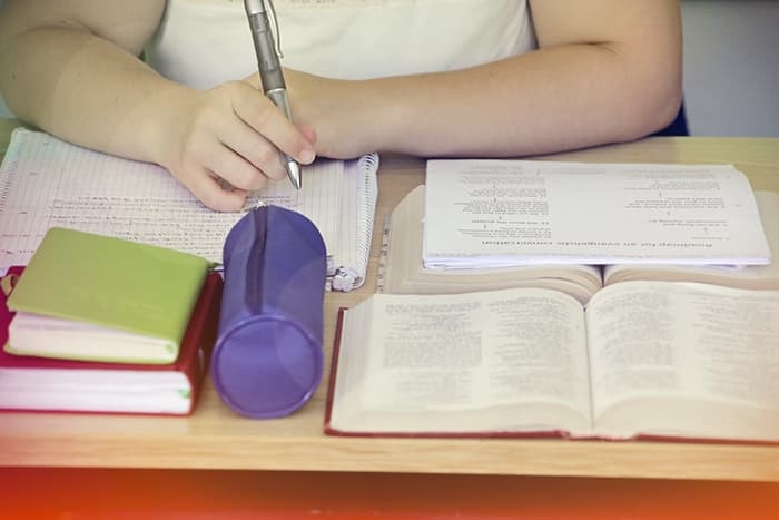 Person, die an einem Schreibtisch mit Büchern, einer Federmappe und aufgeschlagenen Lehrbüchern in ein Notizbuch schreibt.