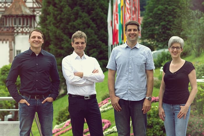 Vier Menschen stehen im Freien, lächelnd, mit Grünzeug und internationalen Flaggen im Hintergrund.