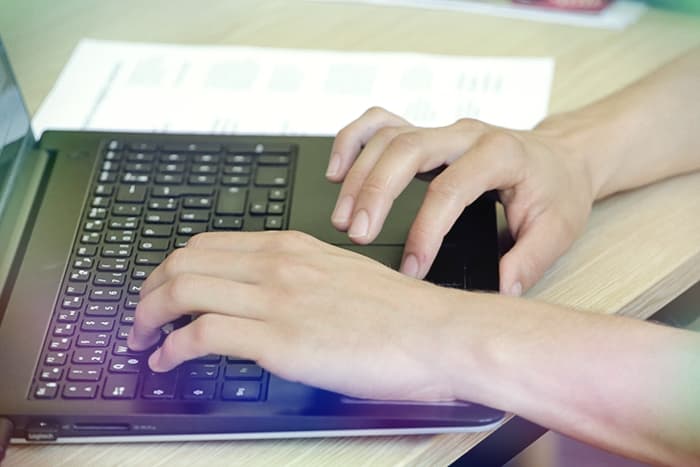 Die Hände tippen auf der Tastatur eines Laptops, während ein Dokument auf dem Schreibtisch neben dem Computer liegt.