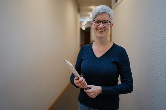 Lächelnde Person mit kurzen grauen Haaren, die ein Klemmbrett in der Hand hält, steht in einem Flur mit cremefarbenen Wänden.