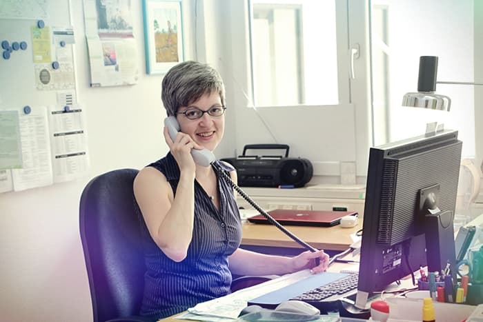 Eine lächelnde Frau mit kurzen Haaren telefoniert an ihrem Bürotisch mit Blick auf einen Computerbildschirm.