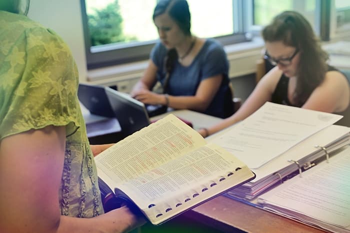 Drei Personen, die an einem Tisch mit Laptops, Papieren und einem offenen Buch in einem hellen Raum mit großen Fenstern lernen.