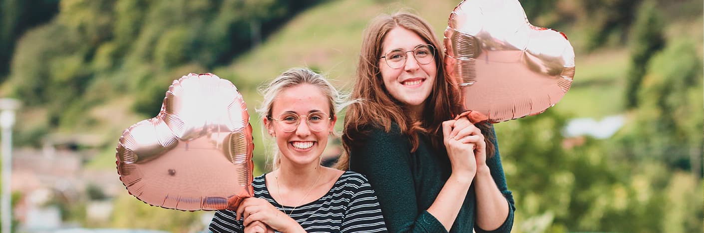 Zwei lächelnde Frauen halten herzförmige Luftballons im Freien mit Grün im Hintergrund.