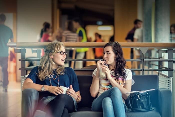 Zwei Frauen sitzen auf einer Couch in einem Haus, unterhalten sich und trinken Getränke, während im Hintergrund Leute spazieren gehen.