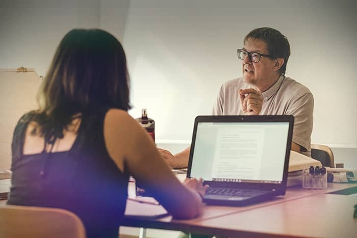 Zwei Personen sitzen an einem Schreibtisch; eine spricht, während die andere mit einem geöffneten Laptop vor sich zuhört.