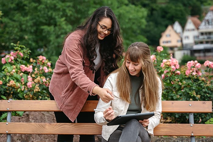 Zwei Frauen lächeln und arbeiten an einem Tablet, während sie auf einer Bank in einem Garten mit rosa Blumen sitzen.