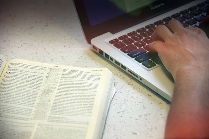 Person, die auf einem Laptop neben einem aufgeschlagenen Buch auf einer hellen Fläche tippt.