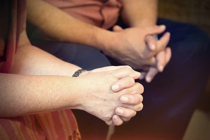 Zwei Personen sitzen mit gefalteten Händen nebeneinander und scheinen sich in einem Moment der Besinnung oder des Gebets zu befinden.